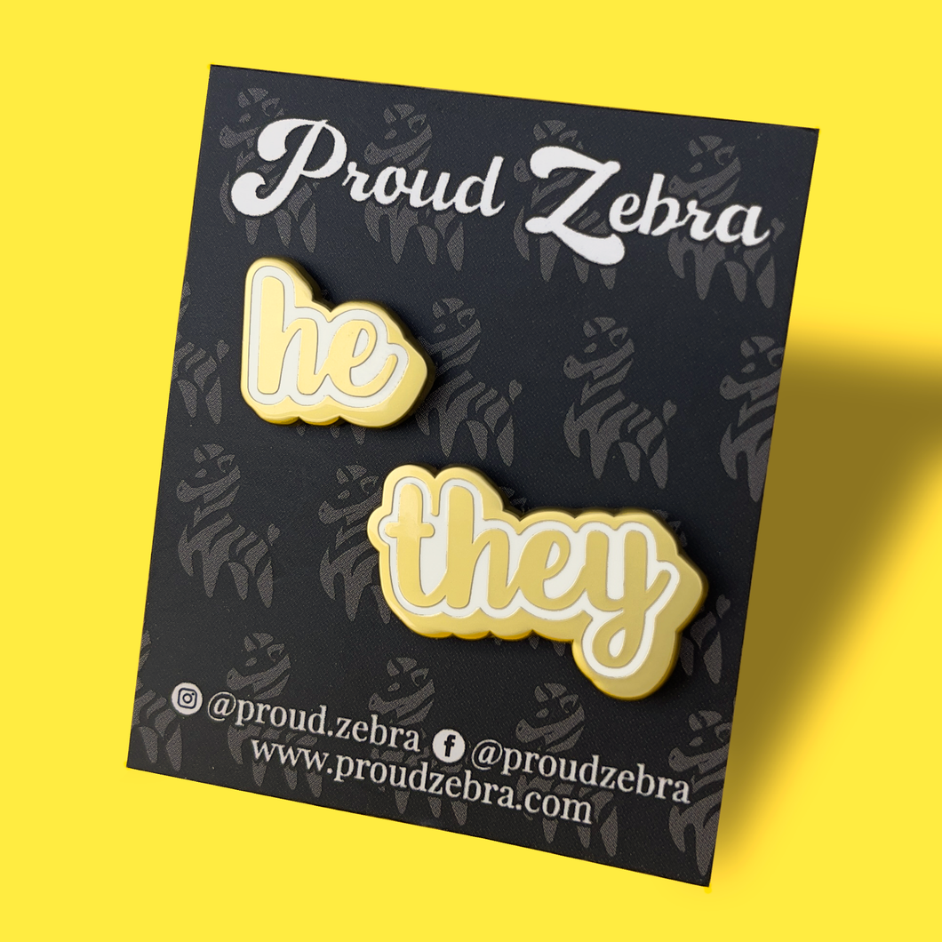 He/They matte gold and white pronoun pins on black backing card by proud zebra