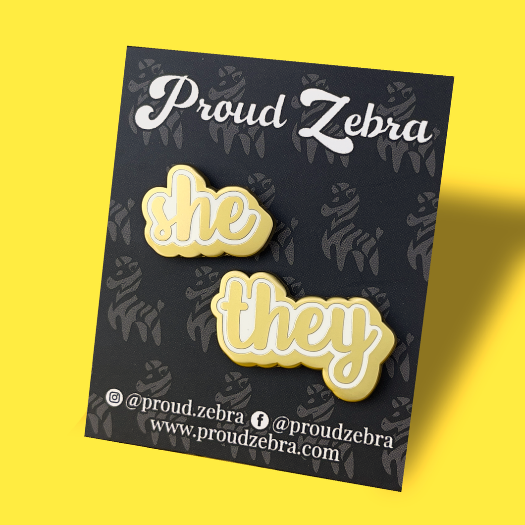 She/They matte gold and white pronoun pins on black backing card by proud zebra