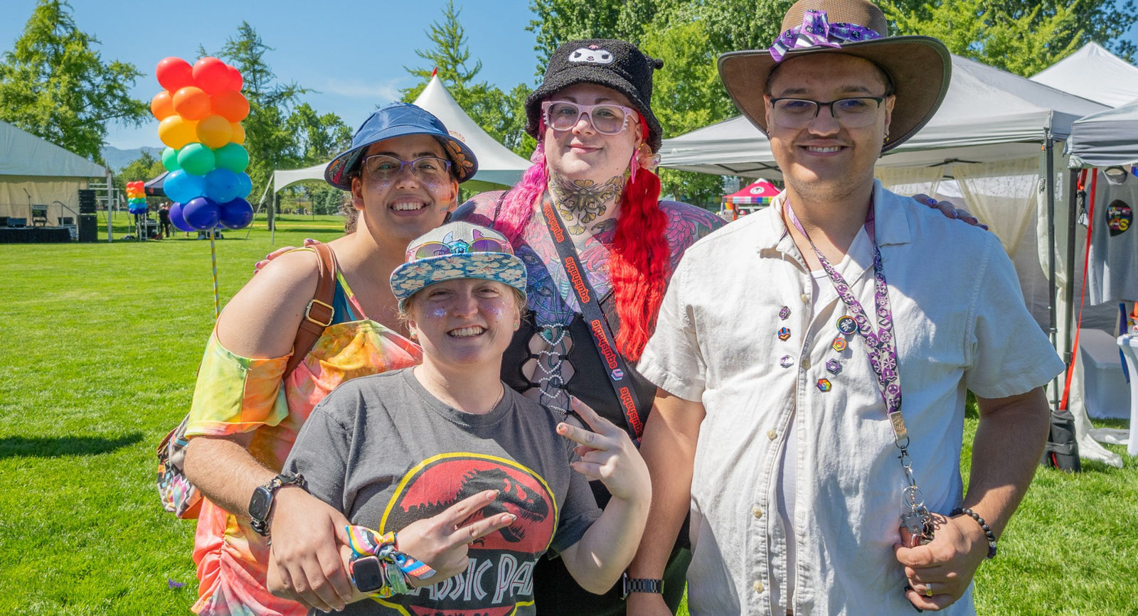 Four smiling LGBTQ+ friends at an outdoor pride festival wearing Proud Zebra enamel pins, lanyards and pride accessories