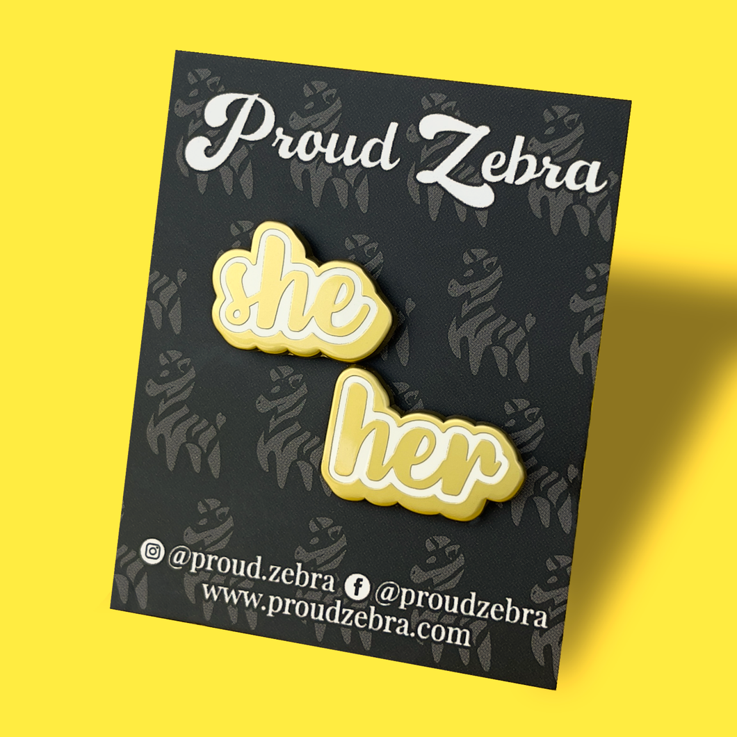 She/her matte gold and white pronoun pins on black backing card by proud zebra